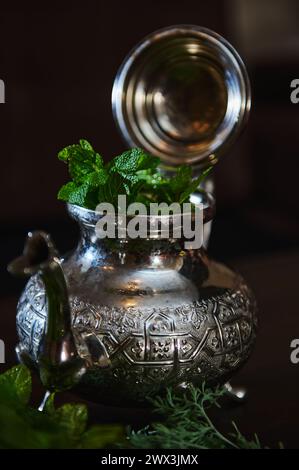 Eine silberne Teekanne und ein Haufen frischer grüner Minze im Vordergrund. Traditioneller marokkanischer Tee als Symbol der Gastfreundschaft der marokkanischen Menschen. Selektiv Stockfoto