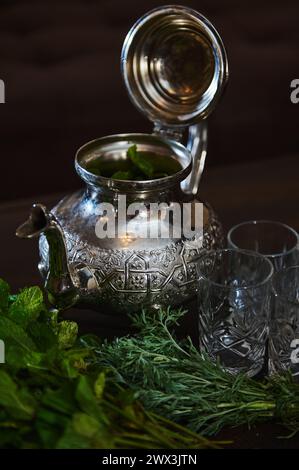 Eine silberne Teekanne und ein Haufen frischer grüner Minze im Vordergrund. Traditioneller marokkanischer Tee als Symbol der Gastfreundschaft der marokkanischen Menschen. Selektiv Stockfoto