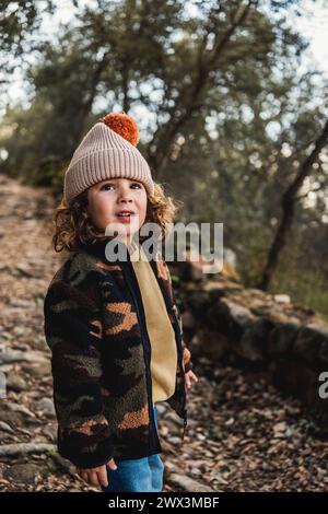 Porträt eines hübschen, modischen kleinen Jungen mit Hut, der posiert, während er auf einem Waldweg wandert Stockfoto