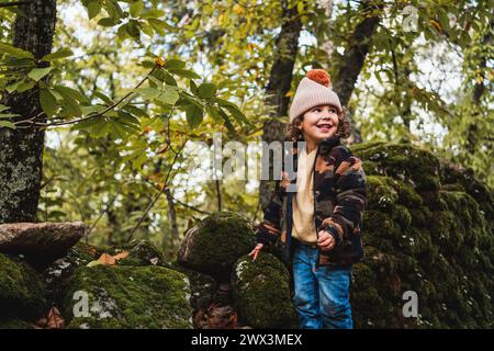 Porträt eines modischen kleinen Jungen mit Hut, der glücklich auf einem Waldweg spielt, während er seinen Eltern etwas über Pflanzen beibringt Stockfoto