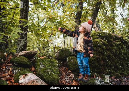 Porträt eines modischen kleinen Jungen mit Hut, der glücklich auf einem Waldweg spielt, während er seinen Eltern etwas über Pflanzen beibringt Stockfoto