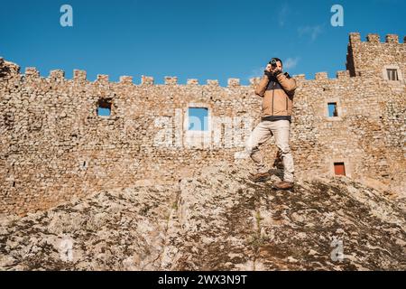 Junger Fotograf auf einem Stein, der Fotos in einer mittelalterlichen Burg macht, Ganzkörperbild aus der Ferne mit Wand im Hintergrund an sonnigen Tagen Stockfoto