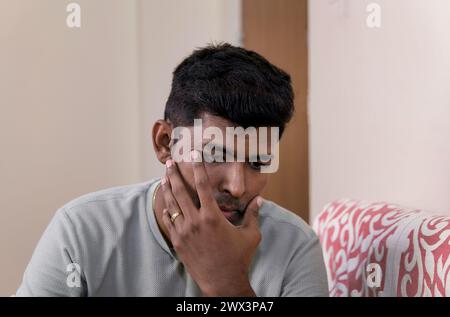 Ein junger Indianer, der zu Hause auf einem Sofa sitzt, wischt eine Träne weg, während er nachdenklich auf den Zuschauer blickt. Sein Ausdruck ist ein Ausdruck von nachdenklicher Traurigkeit Stockfoto