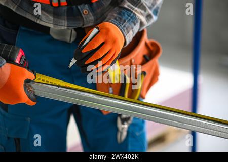 Ein Bauarbeiter in Overall, Handschuhen und einem Werkzeuggürtel misst die Länge eines Gipskartonprofils mit einem Meter – Nahansicht. Stockfoto