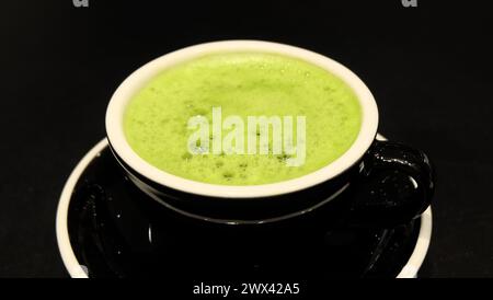 Nahaufnahme einer Tasse Matcha-Grüntee-Drink, mit etwas Schaum auf der Oberseite. Mit einer schwarzen Tasse und einem schwarzen Hintergrund. Stockfoto