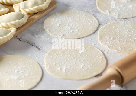 Eingekreister Teig für die Herstellung argentinischer Empanadas auf weißem Marmorhintergrund, eine Nudelnadel und einige ungekochte Empanadas. Stockfoto