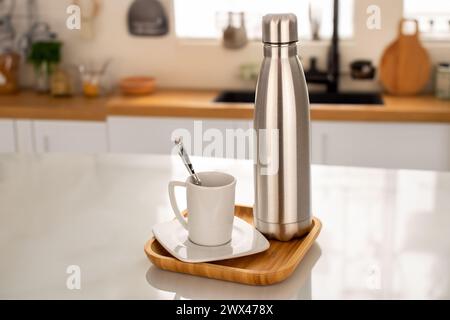 Eine Wasser- oder Kaffeeflasche aus Metall mit Vakuumisolierung auf Holztablett und einer weißen Tasse auf dem Küchentisch. Stockfoto