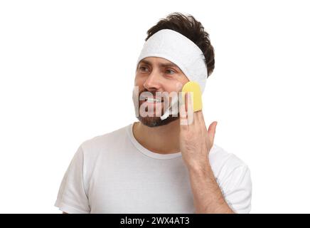 Mann mit Stirnband, der sein Gesicht mit Schwamm auf weißem Hintergrund wäscht Stockfoto