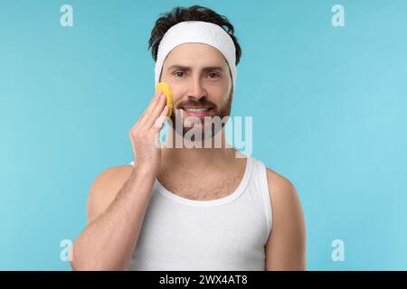 Mann mit Stirnband, der sein Gesicht mit Schwamm auf hellblauem Hintergrund wäscht Stockfoto