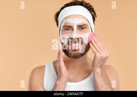 Mann mit Stirnband, der sein Gesicht mit einem Schwamm auf beigem Hintergrund wäscht Stockfoto