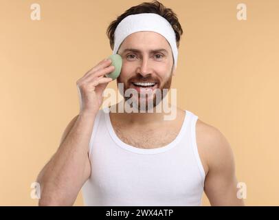 Mann mit Stirnband, der sein Gesicht mit einem Schwamm auf beigem Hintergrund wäscht Stockfoto