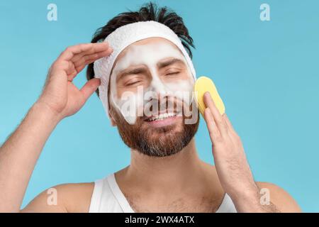 Mann mit Stirnband, der sein Gesicht mit Schwamm auf hellblauem Hintergrund wäscht Stockfoto