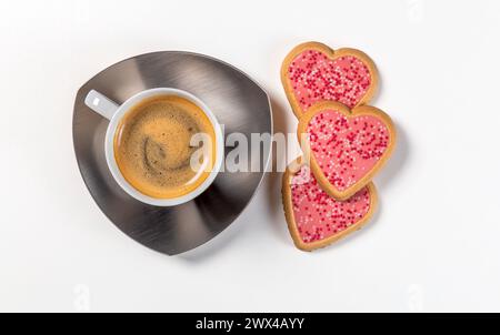 Eine Tasse Kaffee auf einer Untertasse mit drei herzförmigen Keksen in rosa Glasur. Draufsicht auf weißem Hintergrund. Stockfoto