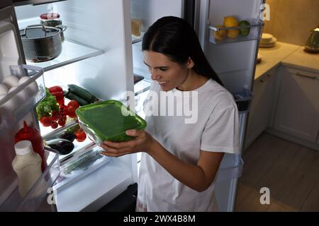 Eine junge Frau, die nachts in der Küche einen Behälter mit Salat aus dem Kühlschrank nimmt Stockfoto