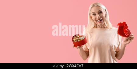 Glückliche reife Frau, die eine Schachtel herzförmiger Schokoladenbonbons auf rosa Hintergrund mit Platz für Text hält Stockfoto