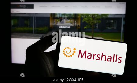 Person mit Smartphone und Logo des britischen Landschaftsprodukteunternehmens Marshalls plc vor der Website. Schwerpunkt auf dem Display des Telefons. Stockfoto