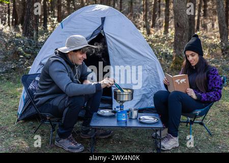 Entspannende Szene auf dem Campingplatz, während ein junges Paar vor ihrem Zelt sitzt. Der Mann bereitet Essen zu, während seine lateinische Freundin gerne ein Buch kreiert Stockfoto