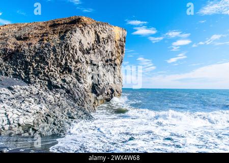 Wellen krachen unter blauem Himmel gegen eine zerklüftete Klippe an der Südküste Islands und zeigen die Schönheit und Kraft der Natur. Stockfoto