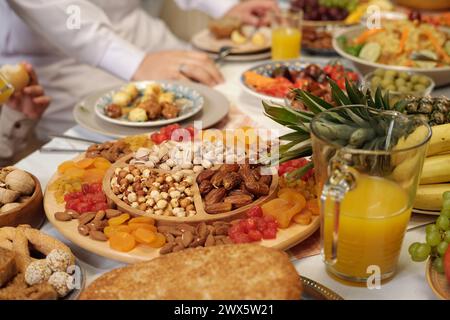 Selektive Fokusaufnahme von süßen getrockneten Früchten und Nüssen auf Holzplatte auf festlichem Esstisch, nicht erkennbare muslimische Menschen feiern Eid Al-Fitr Stockfoto
