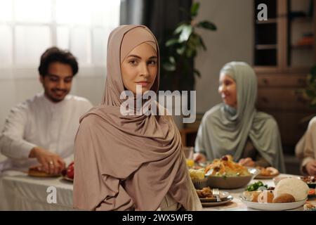 Porträt einer jungen muslimischen Frau, die Hijab trägt, die vor festlichem Tisch sitzt und beim Eid Al-Fitr-Festessen in die Kamera schaut Stockfoto