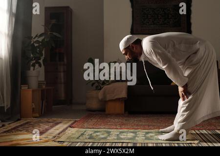 Seitenansicht eines bärtigen muslimischen Mannes, der auf einem Gebetsteg steht und sich nach vorne beugt, während er im Wohnzimmer zu Hause Namaz macht Stockfoto