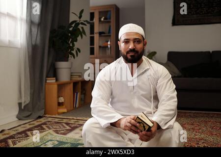 Porträt eines bärtigen muslimischen Mannes, der auf dem Boden im Wohnzimmer sitzt, mit Koranbuch in den Händen, Blick auf die Kamera, Kopierraum Stockfoto