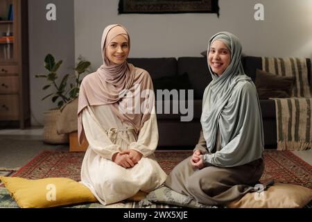 Zwei glückliche junge muslimische Frauen in Hijabs sitzen auf dem Boden im Wohnzimmer und lächeln in die Kamera, mittellanges Porträt Stockfoto