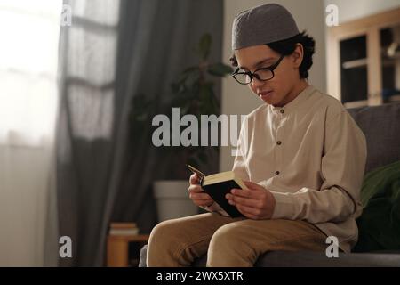 Mittelgroße Aufnahme eines muslimischen Jungen mit Brille, der auf der Couch sitzt und heiliges Koranbuch liest, Kopierraum Stockfoto