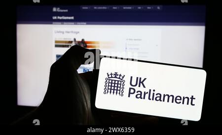 Person, die ein Mobiltelefon mit dem Logo des Legislativgremiums des Vereinigten Königreichs (UK) vor der Webseite hält. Schwerpunkt auf dem Display des Telefons. Stockfoto