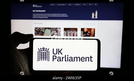 Person, die ein Mobiltelefon mit dem Logo des Parlaments des Vereinigten Königreichs (UK) vor der Webseite hält. Schwerpunkt auf dem Display des Telefons. Stockfoto