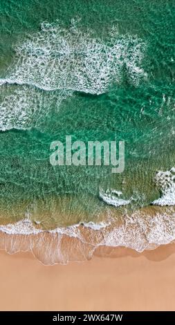 Aus der Vogelperspektive wird die ruhige Schönheit sanfter, transparenter Wellen auf einem Sandstrand sichtbar. Das unberührte Wasser übergeht von Stockfoto