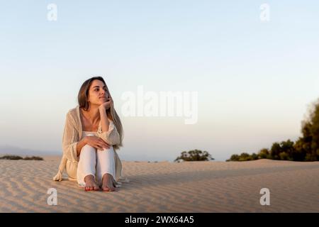 Eine Frau sitzt friedlich auf einer Sanddüne und blickt nachdenklich zum Horizont, während der Sonnenuntergang ein warmes Leuchten auf sie und die Umgebung wirft Stockfoto