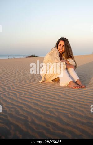 Eine Frau sitzt während des Sonnenuntergangs im weichen Sand der weitläufigen Dünen, ihr Blick richtet sich auf die Kamera. Sie trägt leichte, bequeme Kleidung Stockfoto