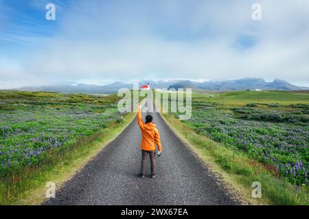 Leistung eines jungen asiatischen Mannes, der auf der Straße mit der heiligen Ingjaldsholskirkja-Kirche auf dem Hügel zwischen Lupinen-Wildblumen steht, die im Sommer in Snaefell blühen Stockfoto