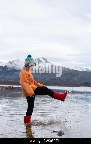 Junge, der mit Wasser in einem See spielt, trägt Gummistiefel mit schneebedeckten Bergen im Hintergrund Stockfoto