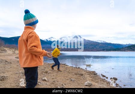 Kinder, die im Winter Steine in einen See werfen, mit schneebedeckten Bergen dahinter Stockfoto