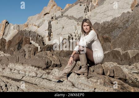 Hübsche blonde Frau sitzt auf felsigem Gelände, trägt ein weißes Kleid und Stiefel, mit zerklüfteten Klippen im Hintergrund Stockfoto