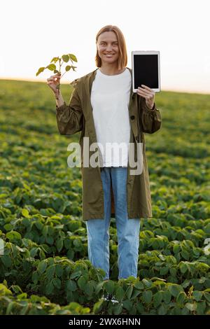 Die lächelnde Agronomin hält ein digitales Tablet mit Mockup-Bildschirm und Sojapflanze im Feld, untersucht, überprüft, schaut in die Kamera. Smart Farming so Stockfoto