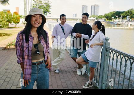 Lächelnde vietnamesische junge Frau, die mit Freunden am Ufer des Flusses rumhängt Stockfoto