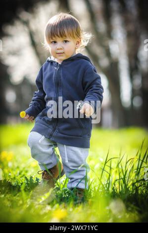 Glücklicher kleiner blonder Junge, der auf einem grünen Feld an bunten Blumen schnüffelt. Niedliches Kleinkind, das die Natur erkundet. Nahaufnahme Stockfoto