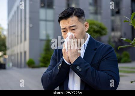 Nahaufnahme eines kranken jungen asiatischen Geschäftsmannes, der auf einer Bank in der Nähe eines Bürogebäudes sitzt und sich die Nase abwischt, mit einer Serviette aus einer laufenden Nase. Stockfoto