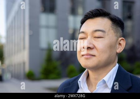 Nahaufnahme eines jungen asiatischen Büroangestellten, der draußen steht, nachdenklich die Augen schließt und sich ausruht. Stockfoto