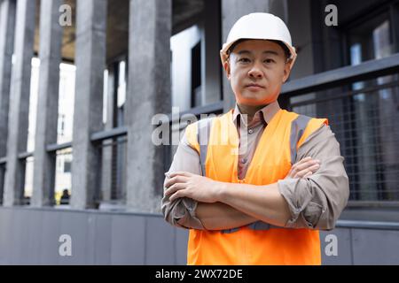 Porträt eines ernsthaften jungen asiatischen Ingenieurarchitekten, der einen Bauhelm und ein Gehäuse trägt, die Hände hebt und selbstbewusst in die Kamera blickt. Stockfoto
