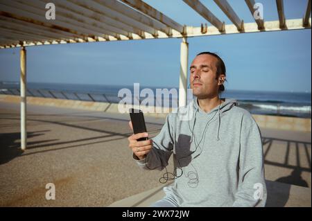 Junger kaukasischer Mann in grauer Sportbekleidung, mit Ohrhörern, Smartphone in der Hand, Musik hören, während er nach dem Workout ruht, auf einer Straße sitzt Stockfoto