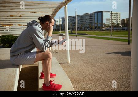 Seitenportrait eines jungen Sportmannes, der Kopfhörer trägt, die mobile App auf seinem Smartphone überprüft, neue Fitness-App durchsucht, während er auf einer Steinbank sitzt Stockfoto