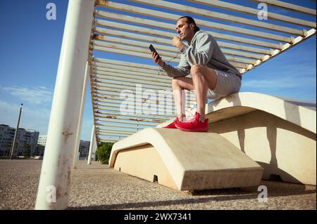 Porträt des jungen, gutaussehenden, sportlichen Mannes in voller Länge, der ein Smartphone benutzt, während er eine Trainingspause auf dem städtischen Sportplatz im Freien macht; Stockfoto