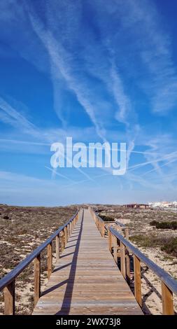 Ein leuchtend blauer Himmel mit weißen Wolken und Flugzeugrauch erstreckt sich über einer langen Holzpromenade auf der Sanddüne von Quiaios Beach. Wunderschöne Landschaft Stockfoto