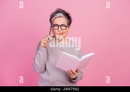 Foto einer pensionierten Sekretärin, die Lehrbuch-Notizbuch hält und Aufgaben auf rosafarbenem Hintergrund löst Stockfoto