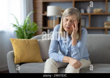 Eine ältere Frau scheint beunruhigt und allein zu sein, sitzt nachdenklich auf einer Couch in einem gut beleuchteten Wohnzimmer und ruft Gefühle der Einsamkeit und Sorge hervor. Stockfoto
