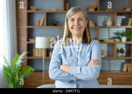 Porträt einer selbstbewussten, Reifen Frau mit einem angenehmen Lächeln, die in einer zwanglosen Home Office-Umgebung mit gekreuzten Armen steht. Stockfoto